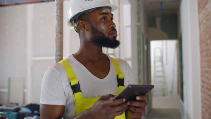African architect inside house being renovated using digital tablet. Portrait of afro-american builder in uniform and helmet checking blueprint on tablet computer