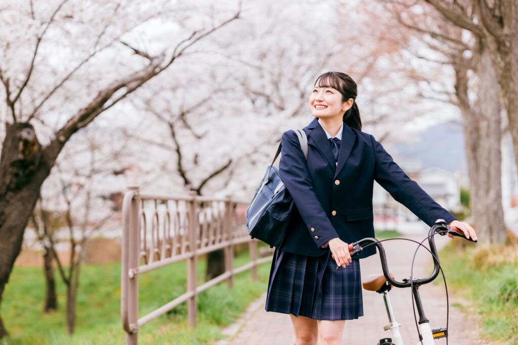 A Japanese high school girl walking along a row of cherry blossom trees while pulling a bicycle.
